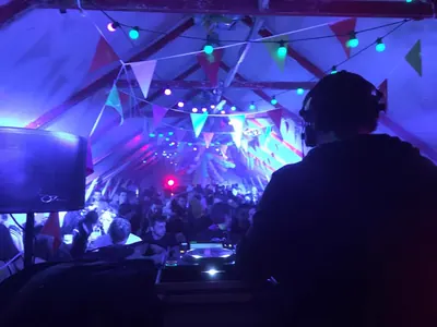 DJ seen from behind under a bunting-draped marquee, crowd in front
