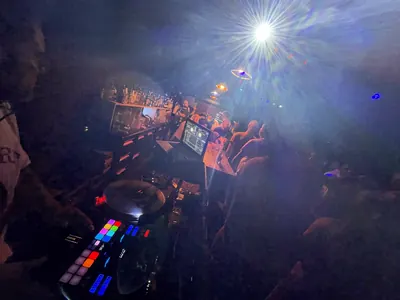 Club DJ booth with lit pads, a spotlight overhead, guests at the bar