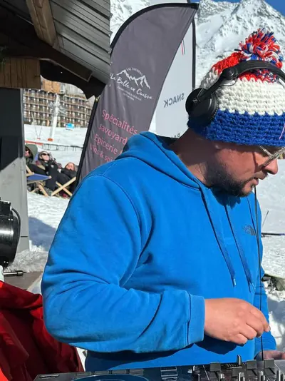 Freddy aux platines en terrasse de station, bonnet et casque, sommets enneigés derrière