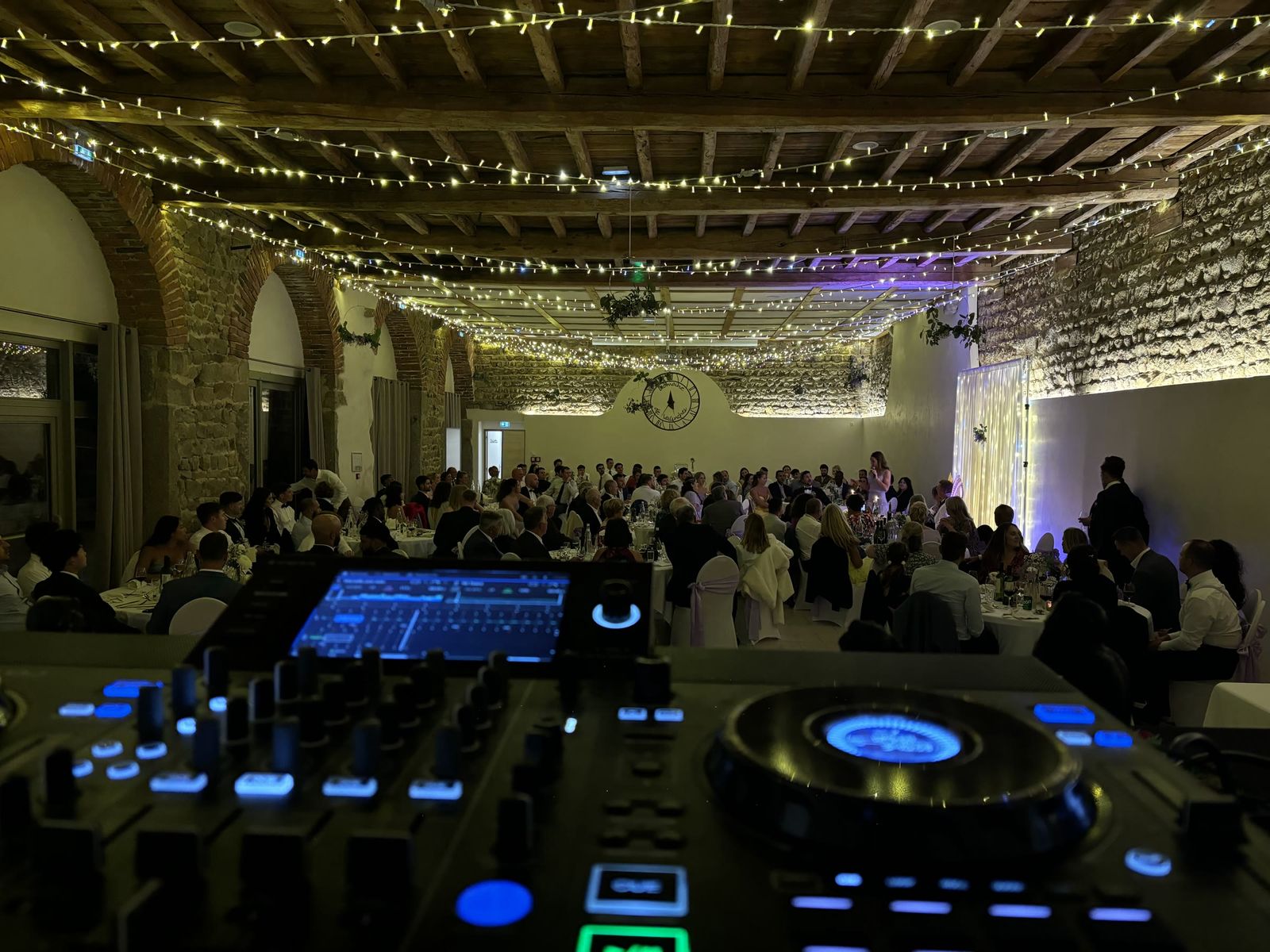 Grande salle de réception aux poutres décorées de guirlandes lumineuses, tables du dîner pleines, vue depuis la régie du DJ