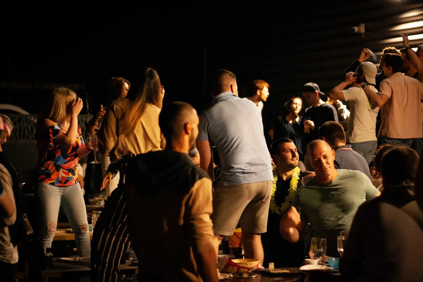 Invités d'une soirée privée qui dansent et chantent autour des tables, de nuit, sous une lumière chaude
